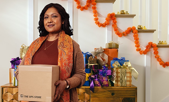 Una mujer sostiene una caja de The UPS Store frente a una mesa llena de regalos.